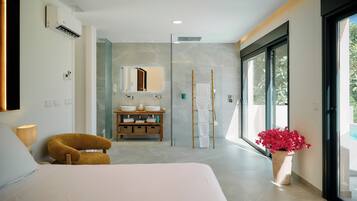 Habitación de hotel moderna con cama, baño con espejo grande, ducha y vista a zonas verdes por las ventanas.