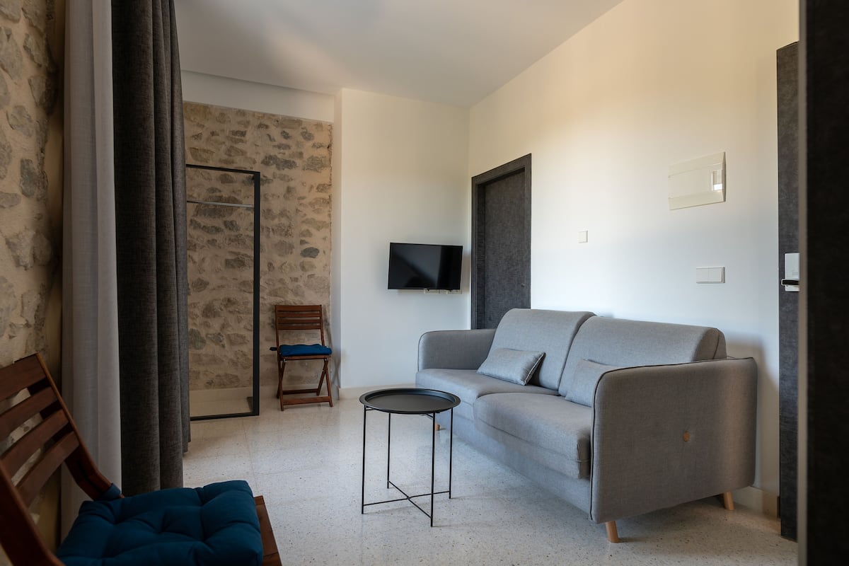 A modern living room with a grey sofa, a small round table, a wooden chair, and a television mounted on the wall.