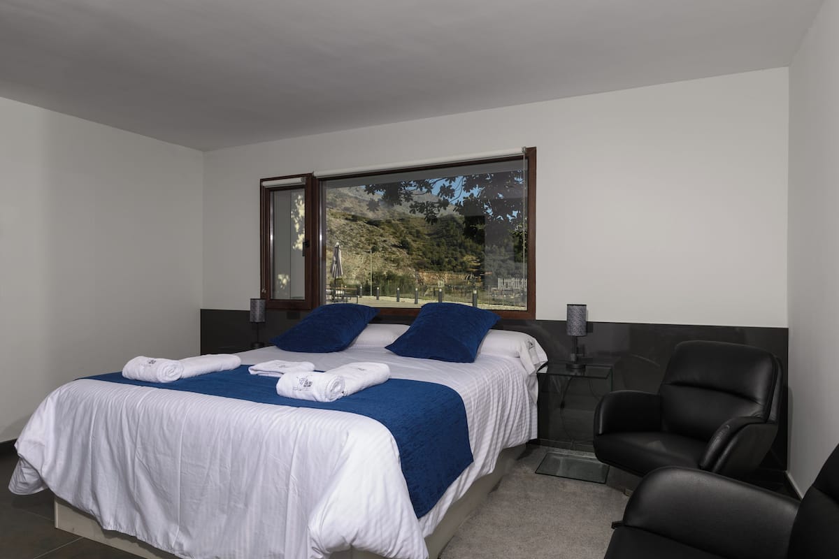A modern hotel room with a large bed, two pillows, a chair, and a window with a view of trees.
