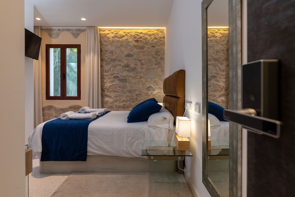 A bedroom with a stone wall, a bed with blue pillows, a glass bedside table, and a TV mounted on the wall.