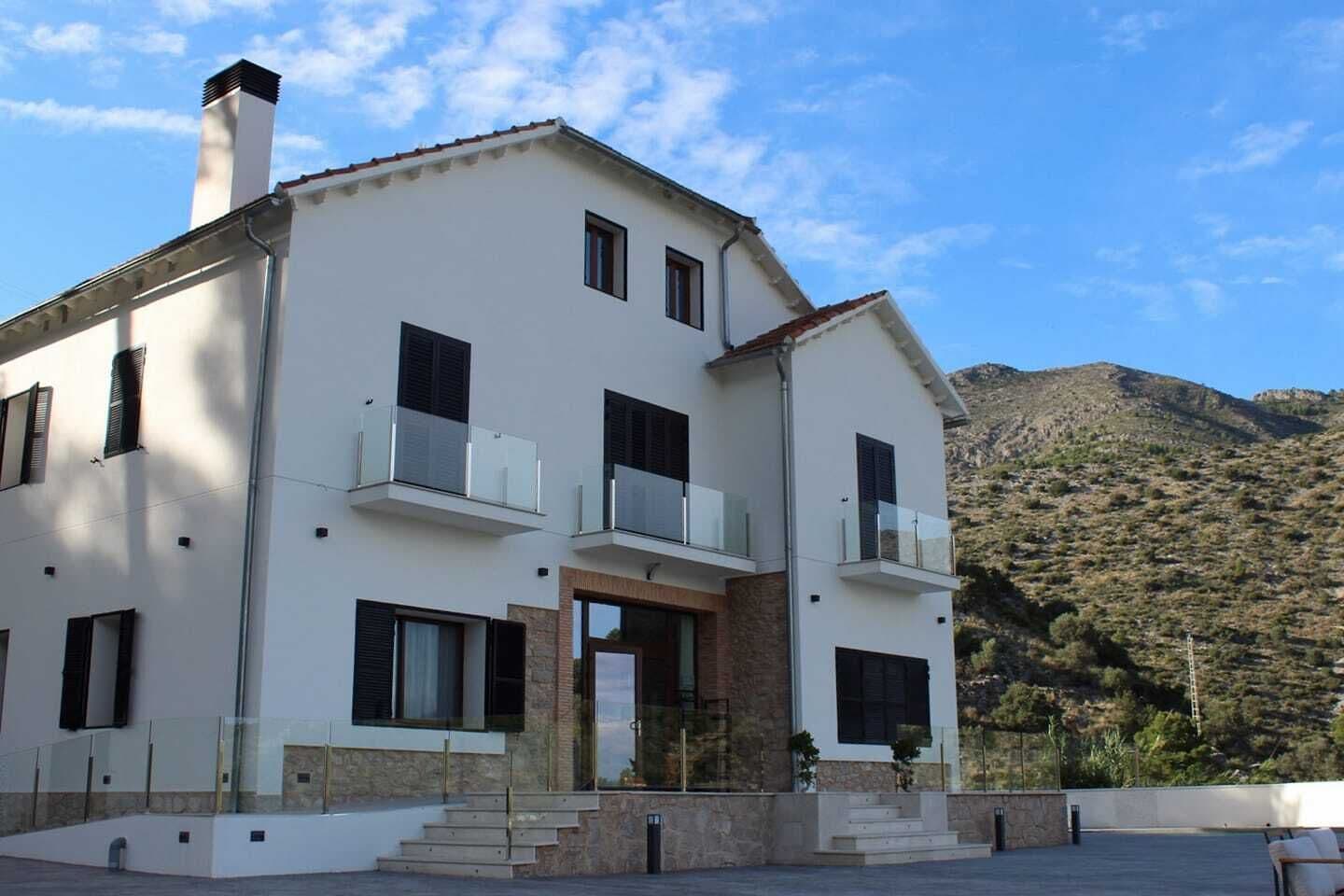 Una casa de dos pisos con fachada blanca, base de piedra y techo de tejas rojas. Cuenta con una entrada principal con puerta de cristal, un balcón en el segundo piso y vistas a un paisaje montañoso.