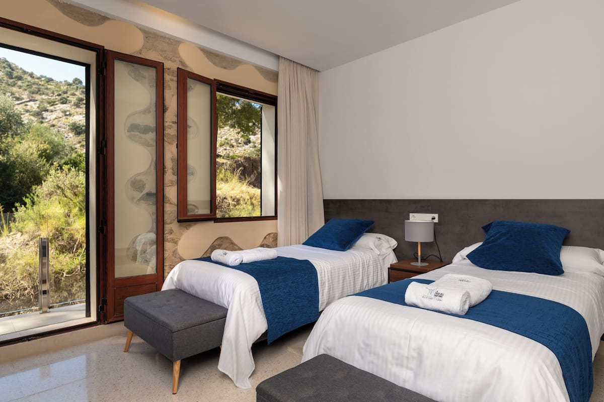 A bedroom with two beds, a large window with a view of the outdoors, and a nightstand with a lamp.