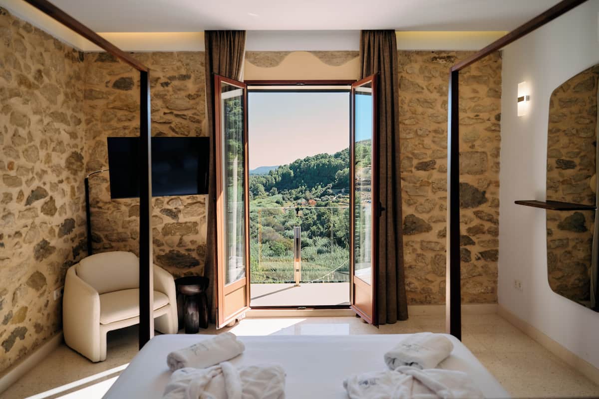 A bedroom with a stone wall, a bed with towels, a chair, a TV, and a balcony with a view of greenery.