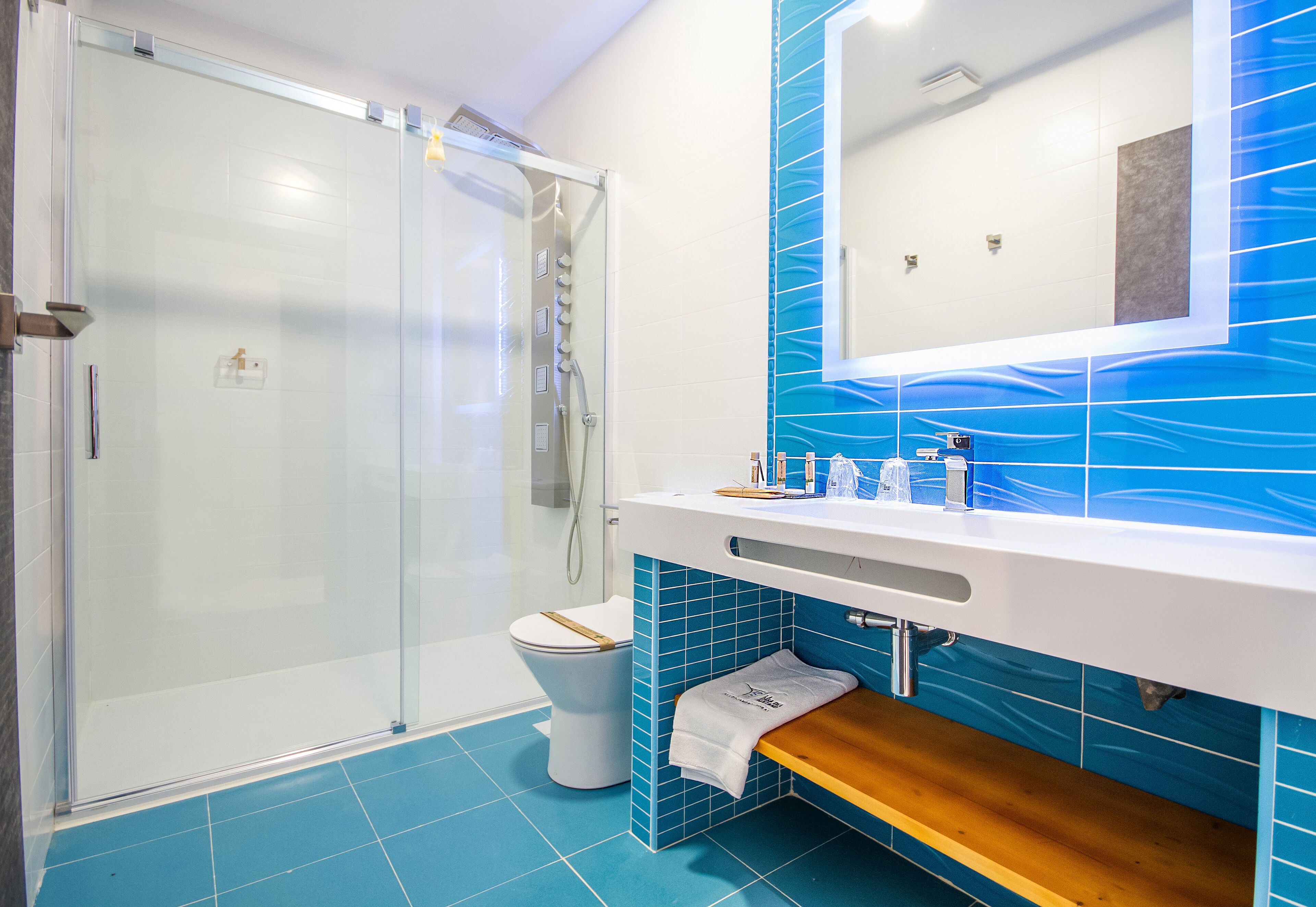 A bathroom with a glass-enclosed shower, a toilet, a sink with a mirror, and blue-tiled walls.