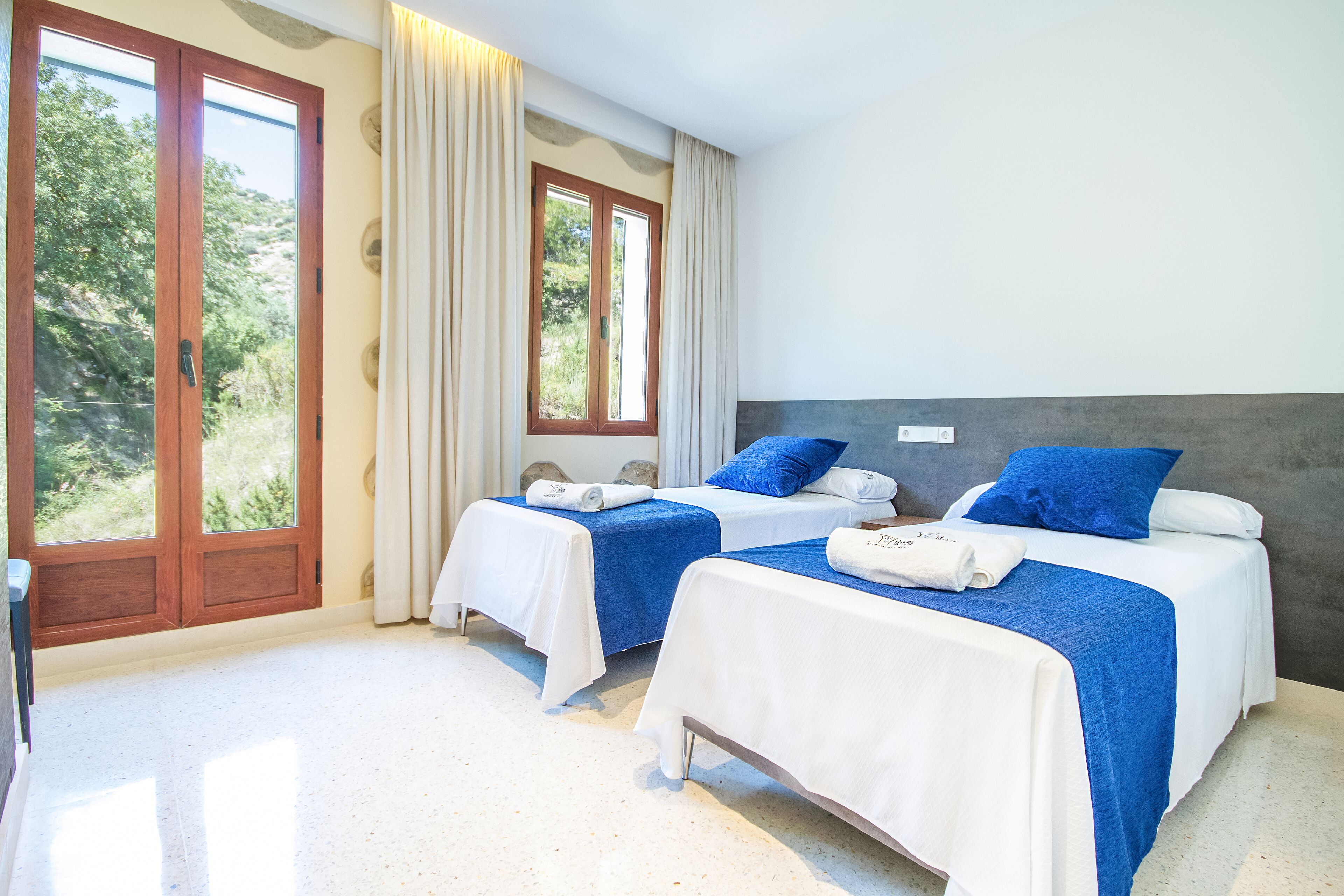 Habitación de hotel con dos camas, cada una con una colcha azul y blanca, y un ventanal grande con cortinas.