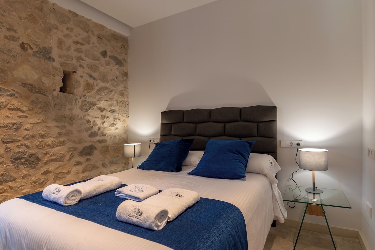 A bedroom with a stone wall, a bed with blue pillows, and two bedside lamps.