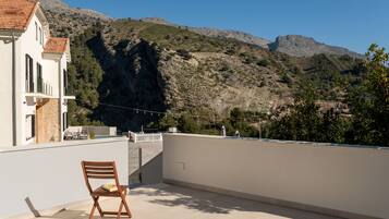 A balcony with a wooden chair, a white building with a tiled roof, and a mountainous landscape in the background.