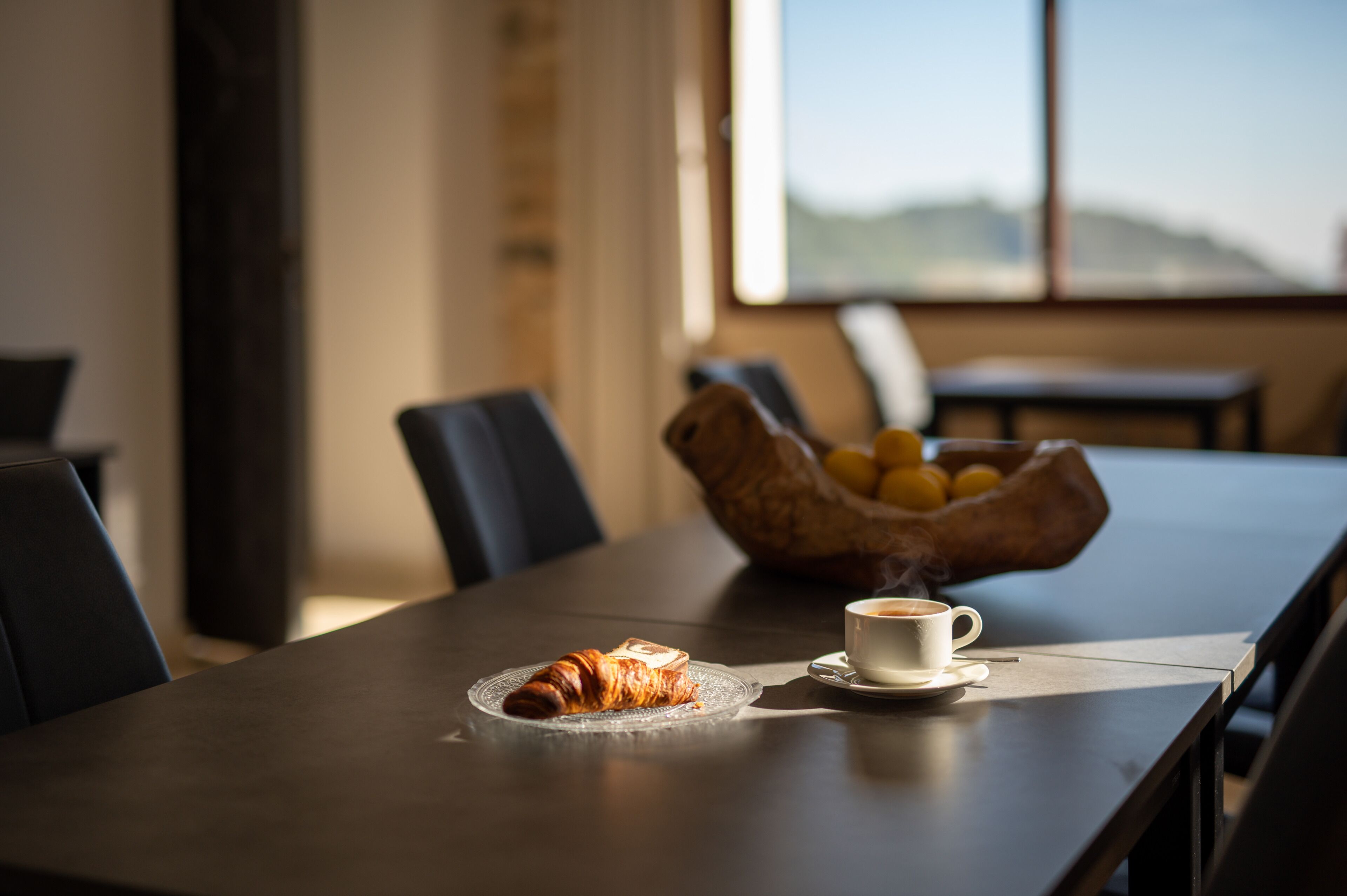 Een eettafel met een croissant, een kop koffie, een schaal fruit en uitzicht op het buiten.