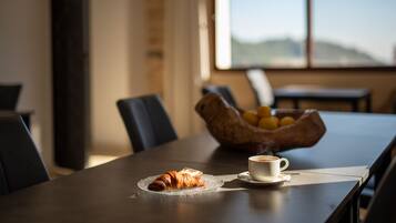 Een eettafel met een croissant, een kop koffie, een schaal fruit en uitzicht op het buiten.