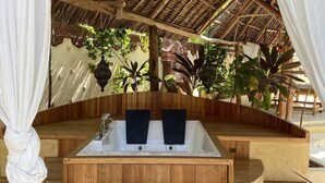 A wooden deck with a hot tub, surrounded by plants and a thatched roof structure.