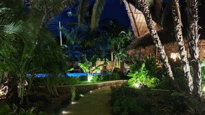 A nighttime view of a tropical resort with a swimming pool, palm trees, and a thatched-roof building.