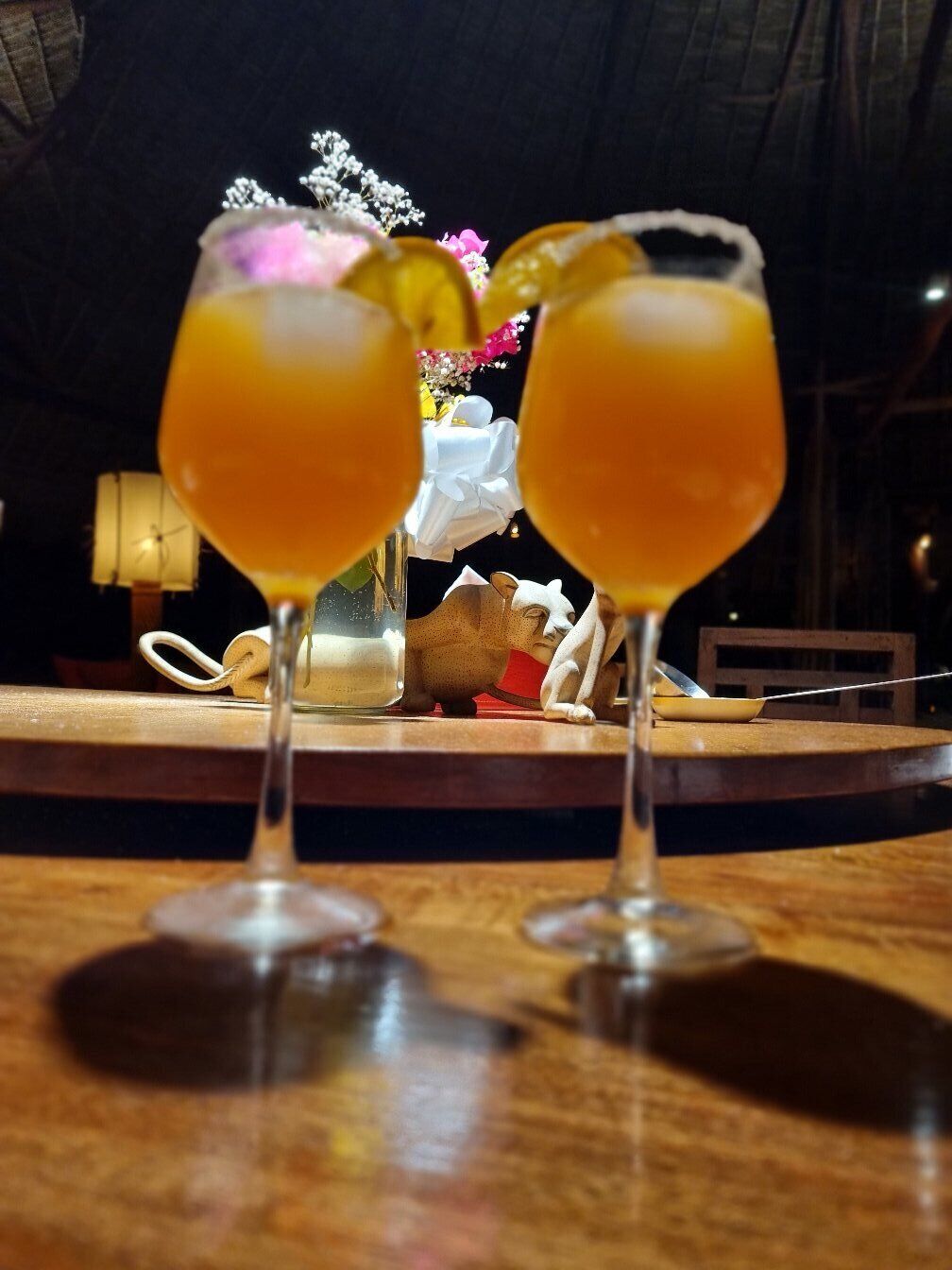 Two glasses of orange beverage on a wooden table with a decorative vase and a small figurine in the background.