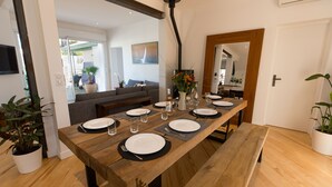 Un espace repas avec une longue table en bois, dressée pour six personnes, entourée de plantes en pot et doté d’un aménagement intérieur moderne.