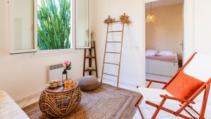A room with a large window, a wicker side table, a wooden ladder, a white chair with an orange cushion, and a bed in the background. - LE FLORIDA AP4340 By Riviera Holiday Homes (Villefranche-sur-Mer)