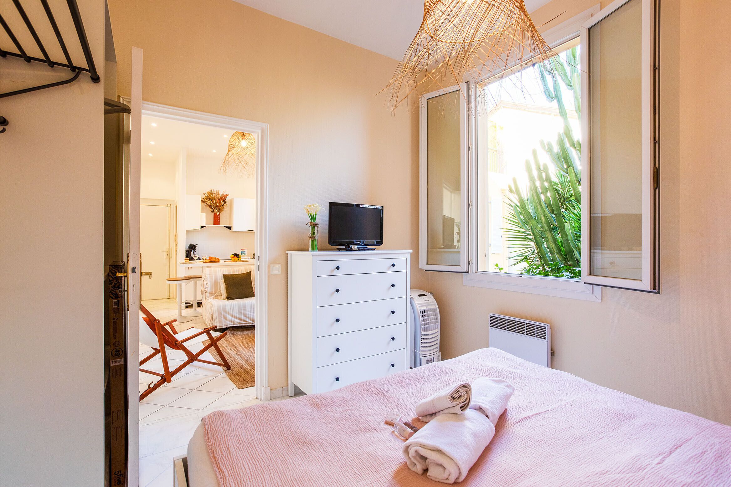 A bedroom with a bed, a dresser, a television, and a window with a view of greenery.