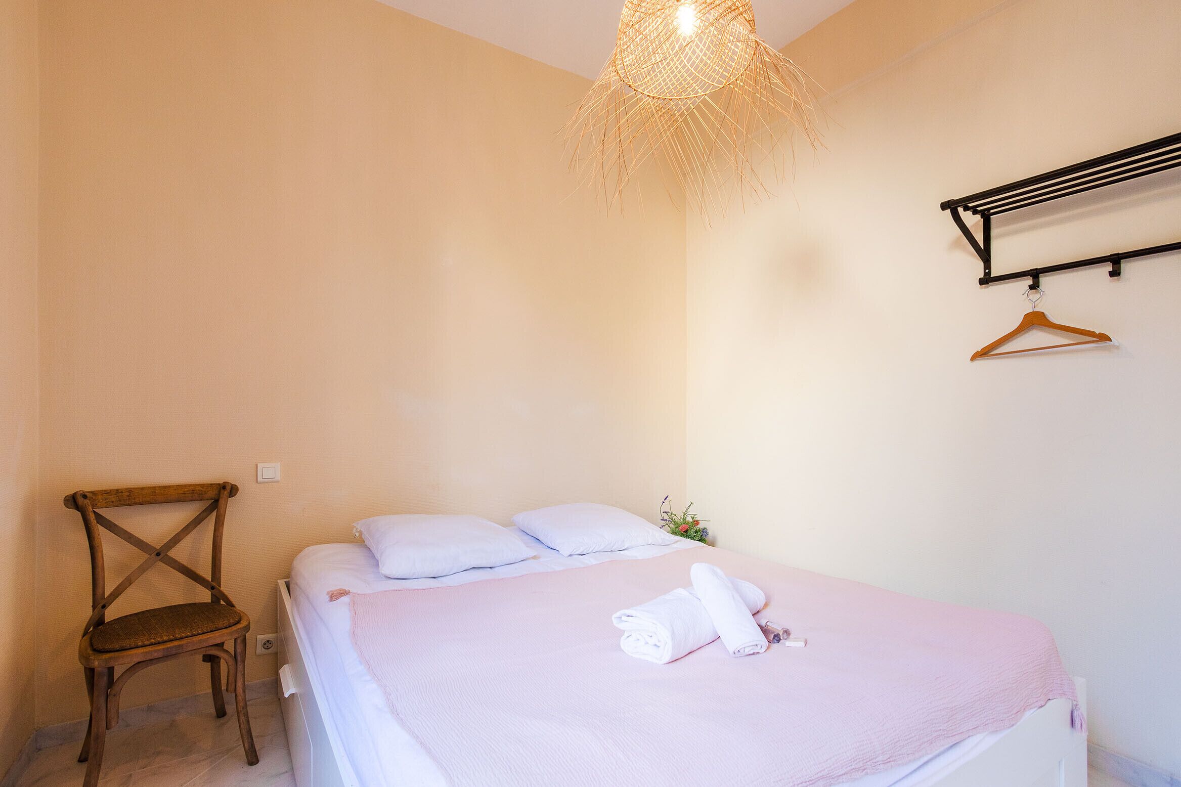 A neatly made bed with a pink blanket and a rolled towel, a wooden chair, a hanging clothes rack, and a unique woven light fixture.