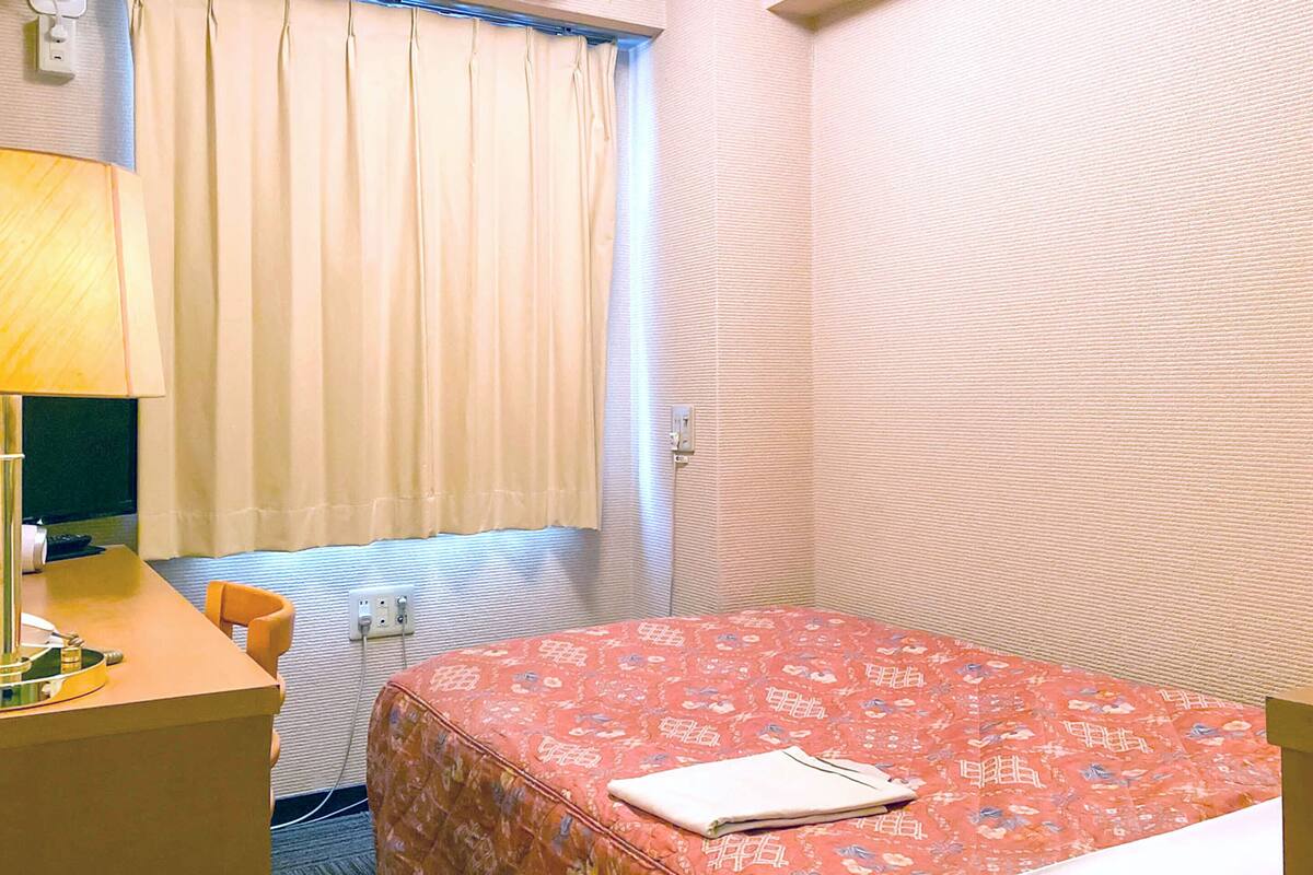 Hotellihuone, jossa on sänky, työpöytä lampun kanssa, tuoli, ikkuna verhoilla ja seinä, jossa on kuvioitu pinta.