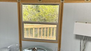 A room with a glass table, chairs, a white lamp, and a window with a view of a wooden deck and trees.