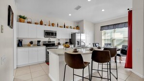 Una cocina moderna con armarios blancos, una isla central con taburetes y un comedor con una cortina roja.