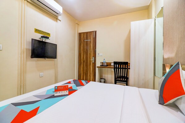 A hotel room with a bed, a television mounted on the wall, a wooden door, a small table with a chair, and a mirror.