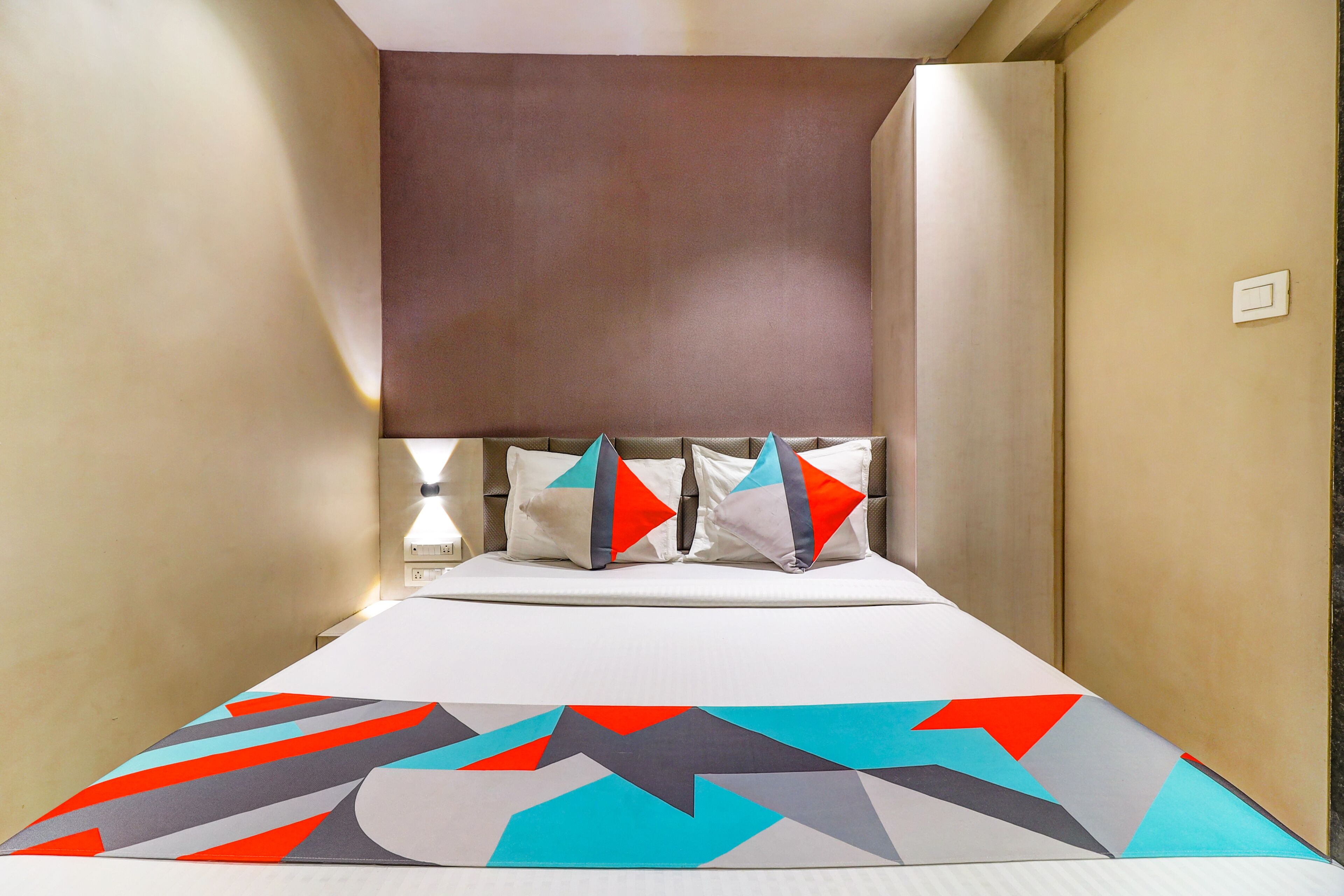 A narrow hotel room with a bed, a headboard, and a bedside table with a lamp.
