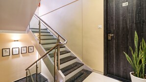 Una escalera con barandilla de cristal que lleva a un rellano, una puerta de madera oscura con placa de número y una planta en maceta en el rellano.