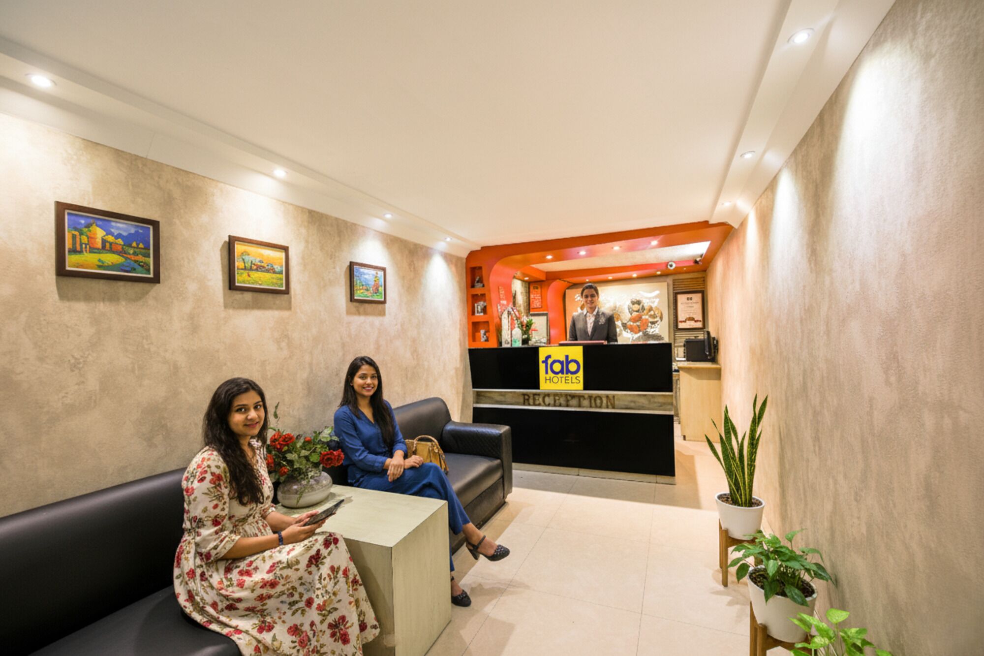 A hotel reception area with a reception desk, two waiting guests, and a few potted plants.