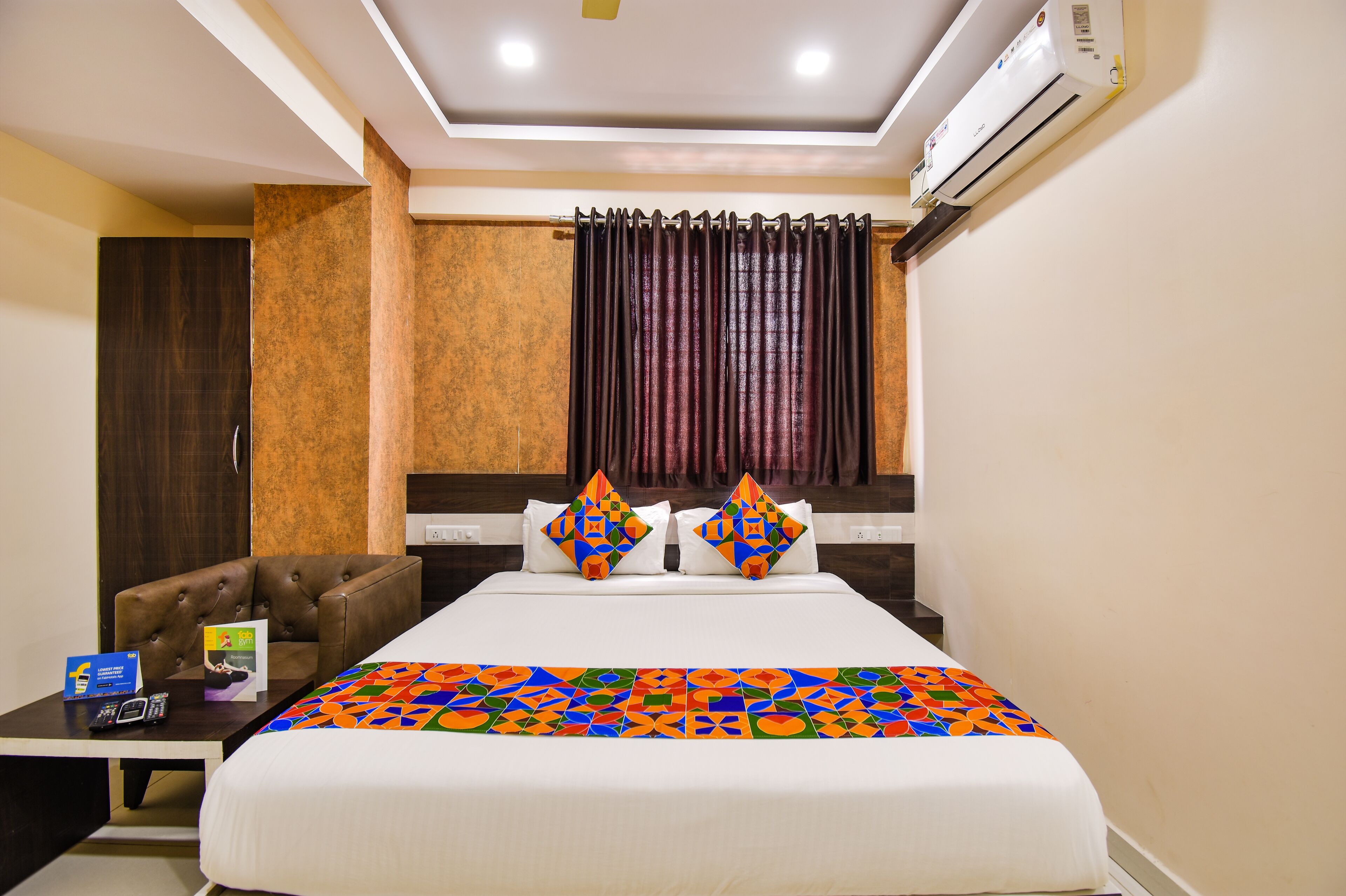 A hotel room with a bed, two decorative pillows, a brown leather chair, a small table with a remote control, and an air conditioning unit.