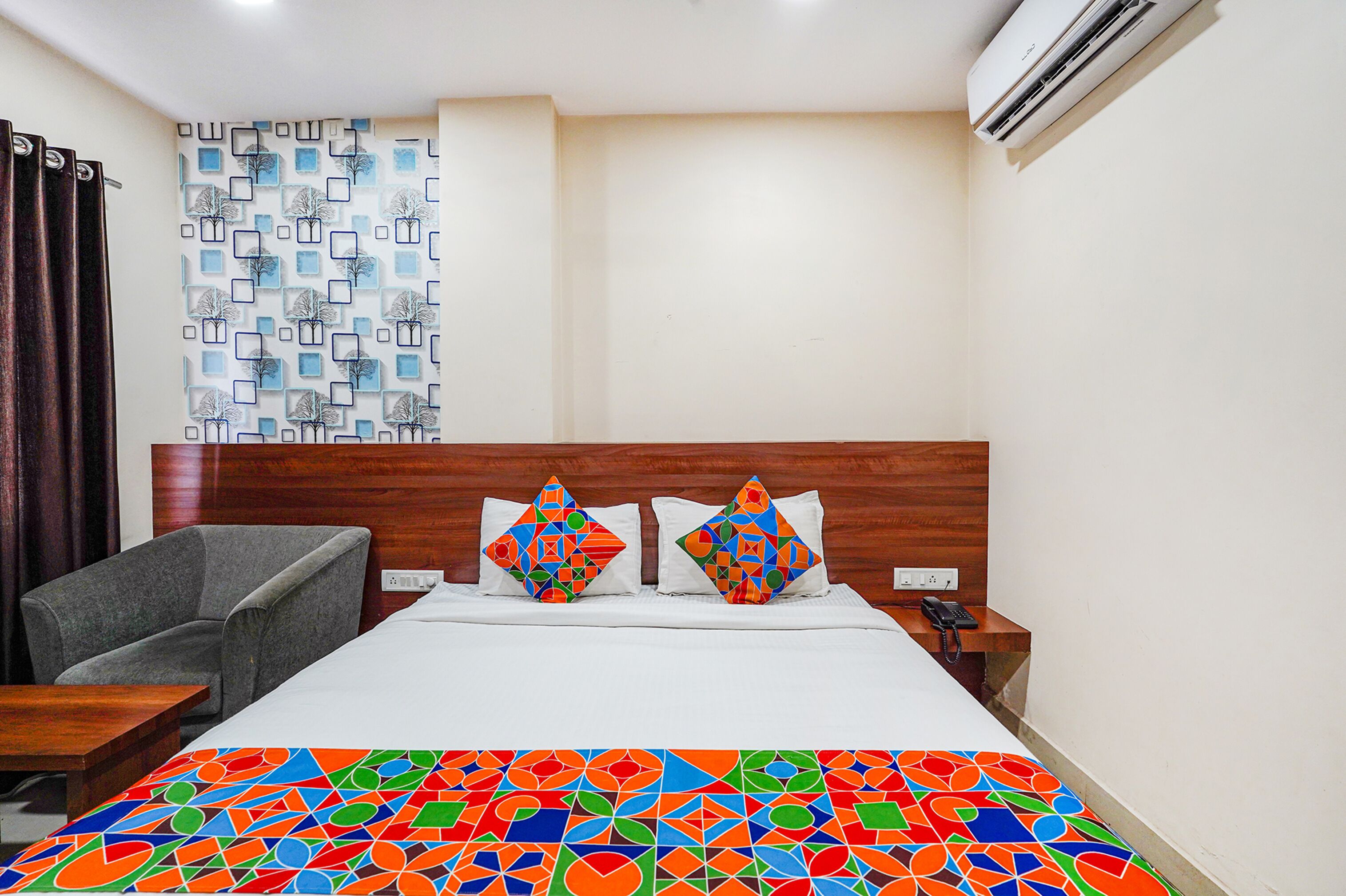 A hotel room with a bed, a chair, a nightstand, and an air conditioning unit.