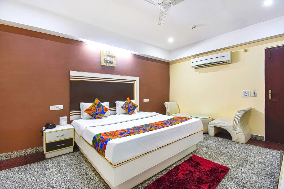 A hotel room with a large bed, a bedside table, a chair, an air conditioning unit, and a red rug.