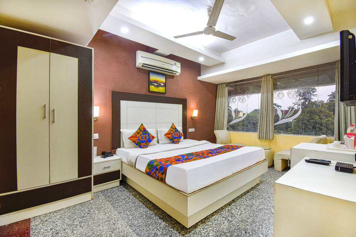 A hotel room with a bed, a desk, a wardrobe, an air conditioner, a ceiling fan, and a window with a view of trees.