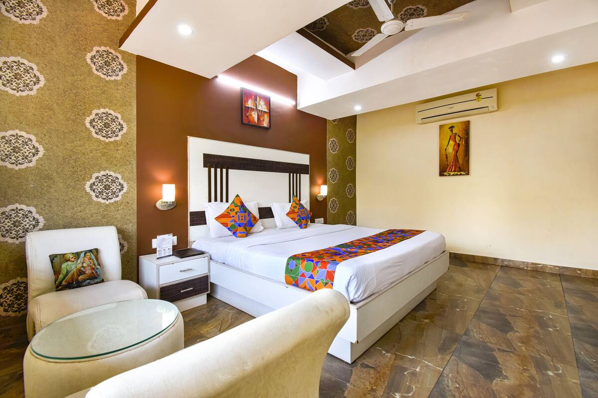 A hotel room with a large bed, a bedside table, a chair, a glass table, patterned walls, and an air conditioning unit.