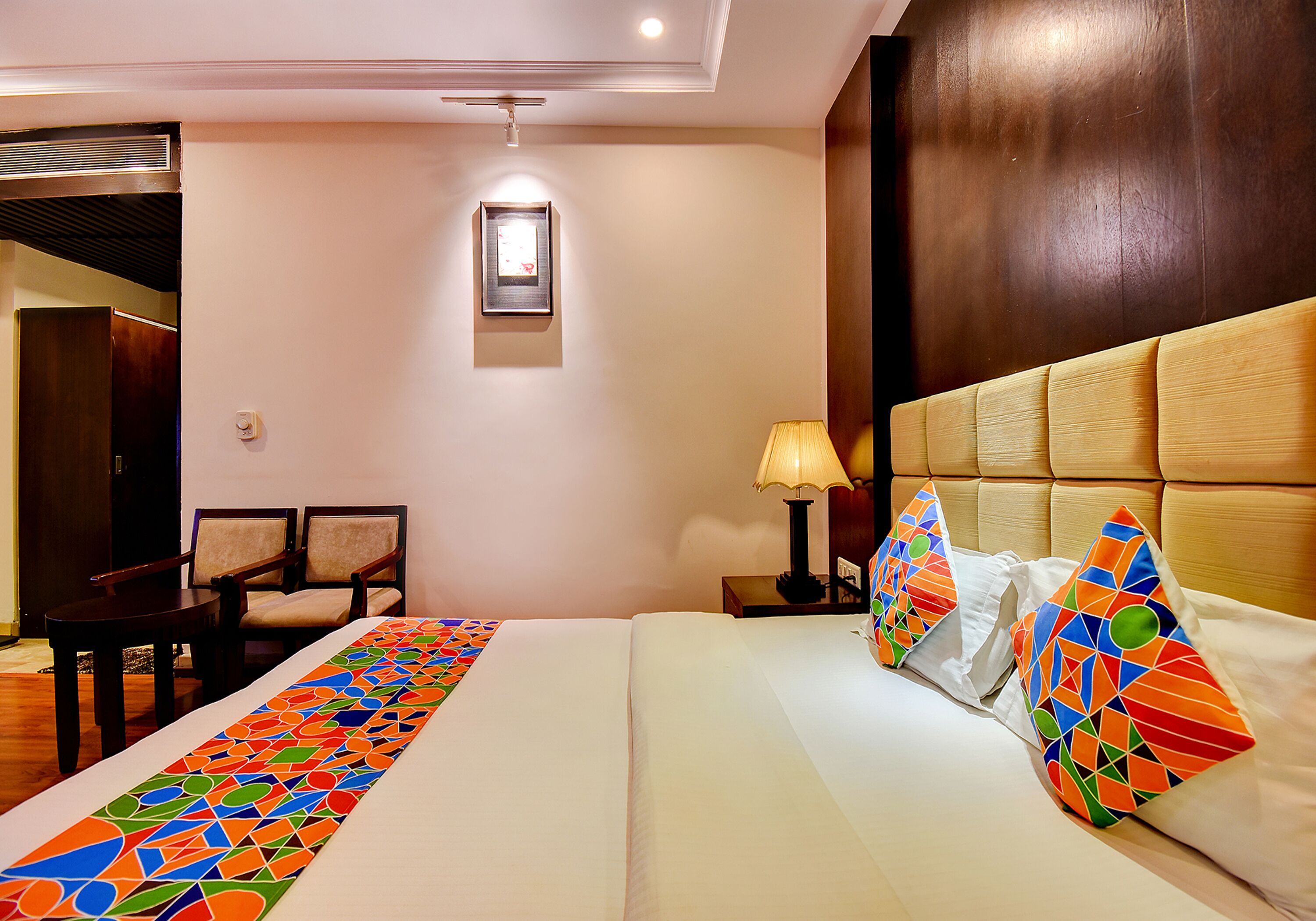 A hotel room with a bed, a desk, two chairs, a nightstand with a lamp, and a framed picture on the wall.