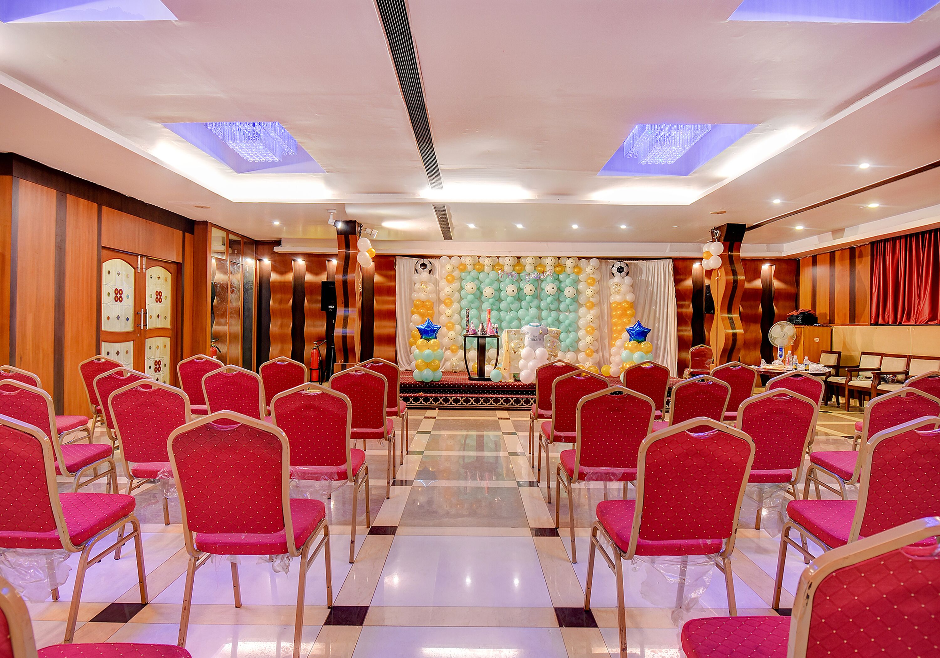 Une salle de conférence avec des chaises rouges disposées en rangées, une scène surmontée d’un décor de ballons multicolores et un sol à carreaux.