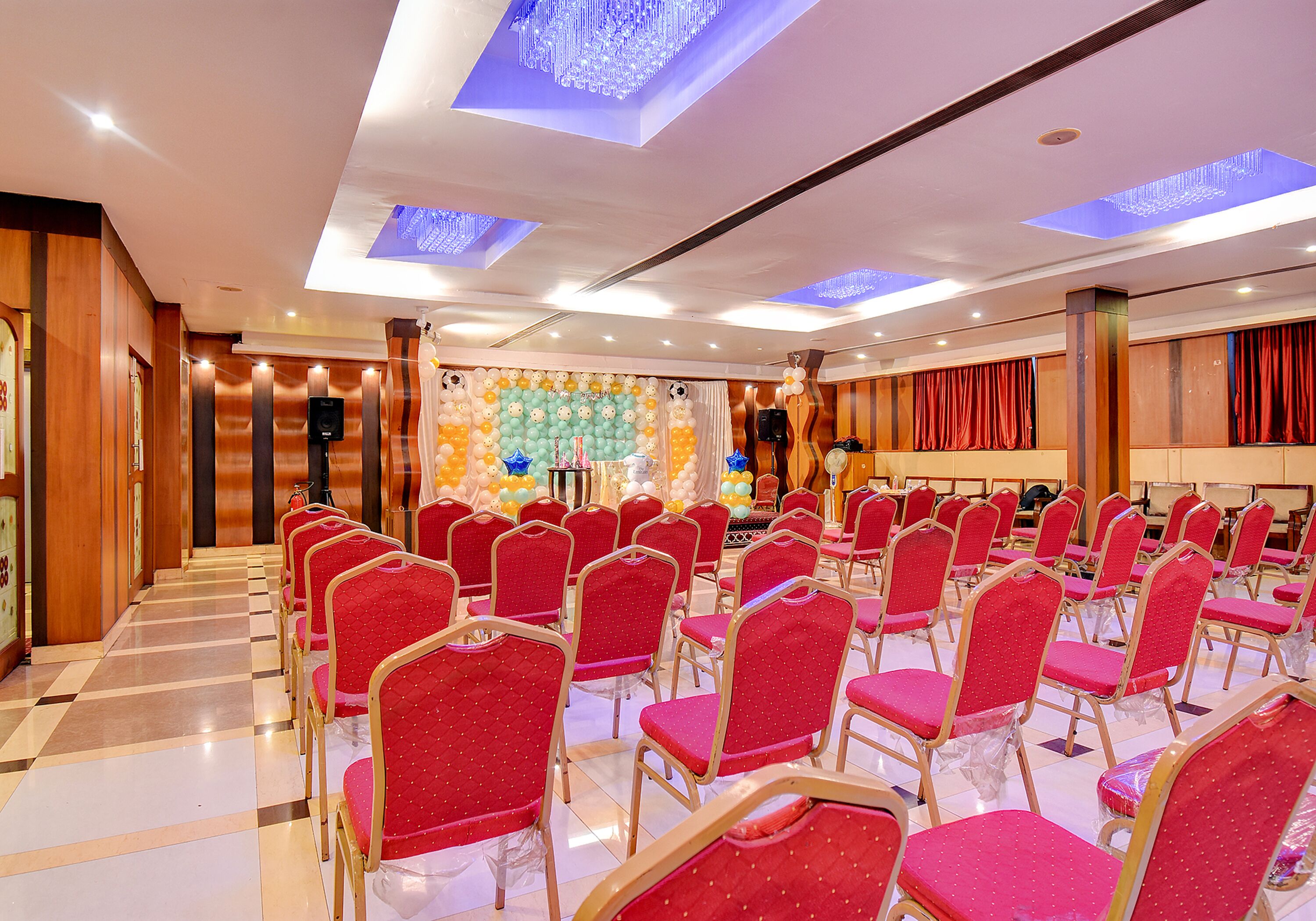 Un salón de conferencias con filas de sillas rojas, un escenario con un orador y una pared decorada con globos.
