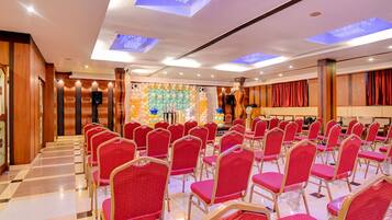 Un salón de conferencias con filas de sillas rojas, un escenario con un orador y una pared decorada con globos.