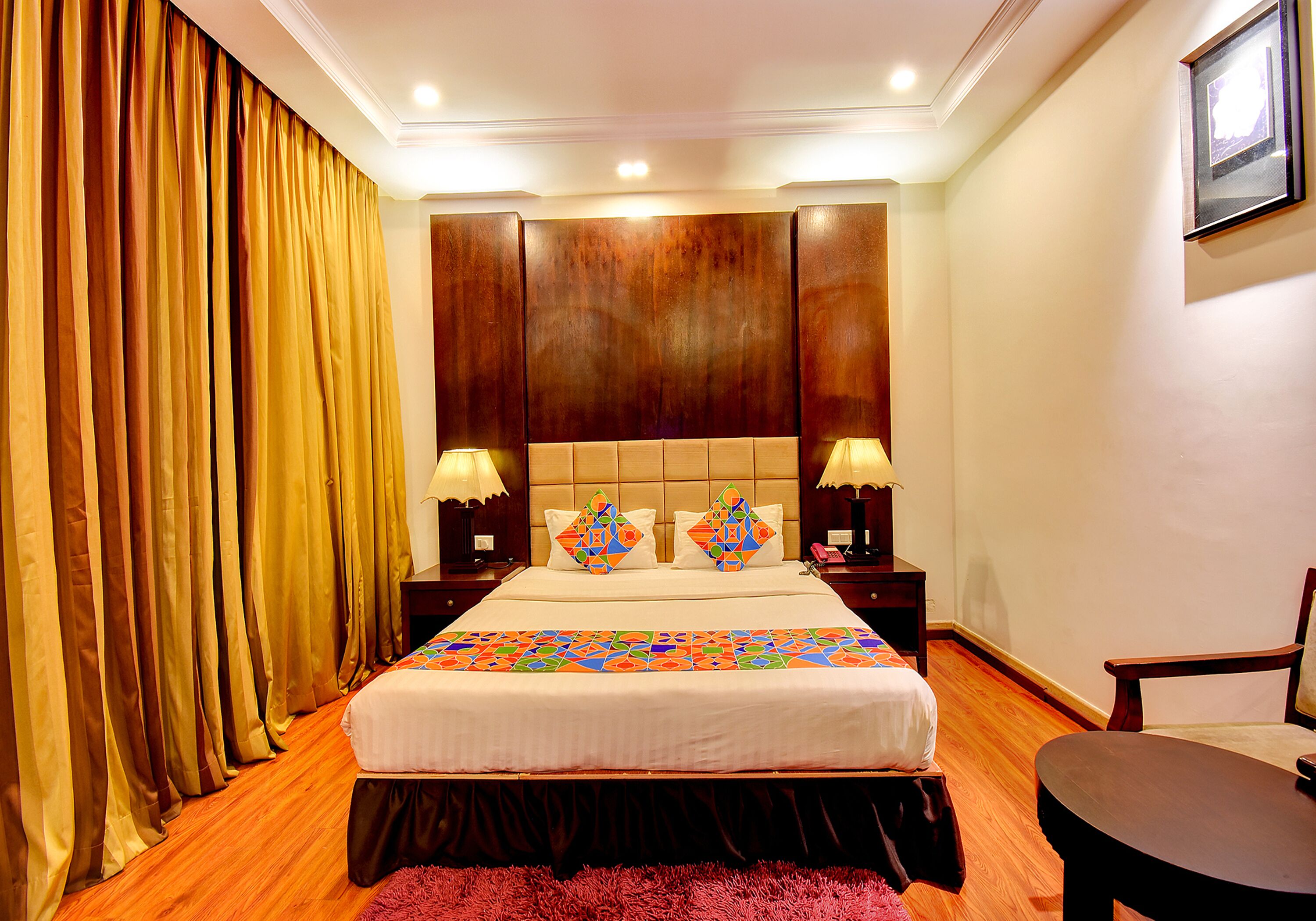 Habitación de hotel con una cama grande, cabecero de madera, dos mesitas de noche con lámparas, una silla y un ventanal con cortinas.