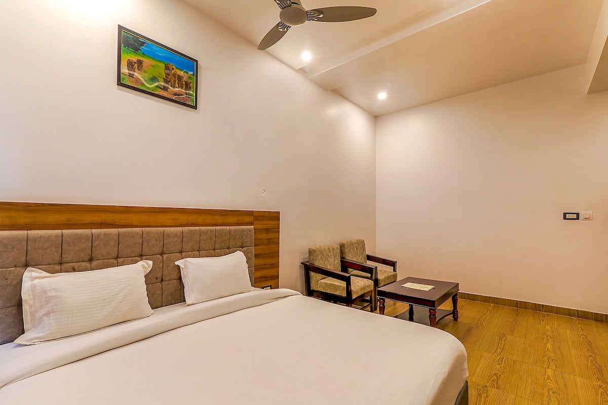 A hotel room with a bed, two chairs, a small table, a ceiling fan, and a painting on the wall.
