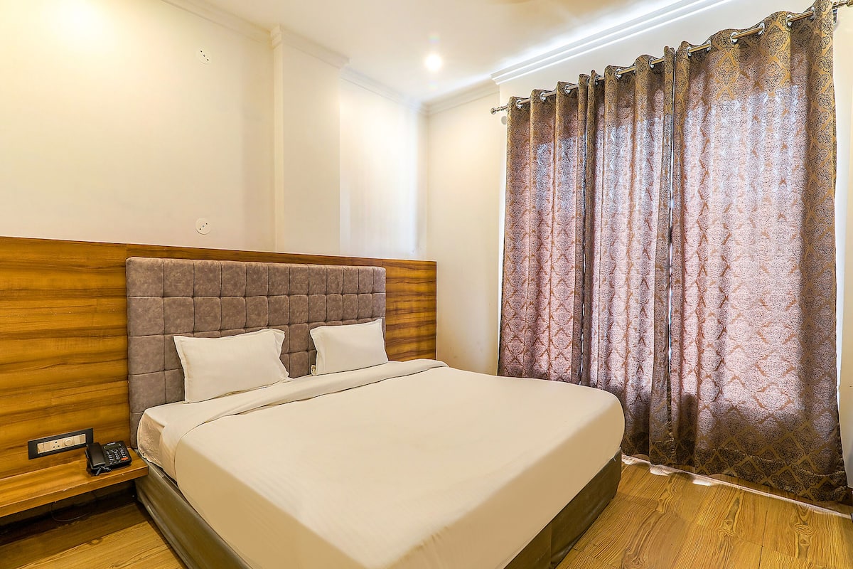 A neatly made bed with a wooden headboard, a bedside telephone, and a window with patterned curtains.