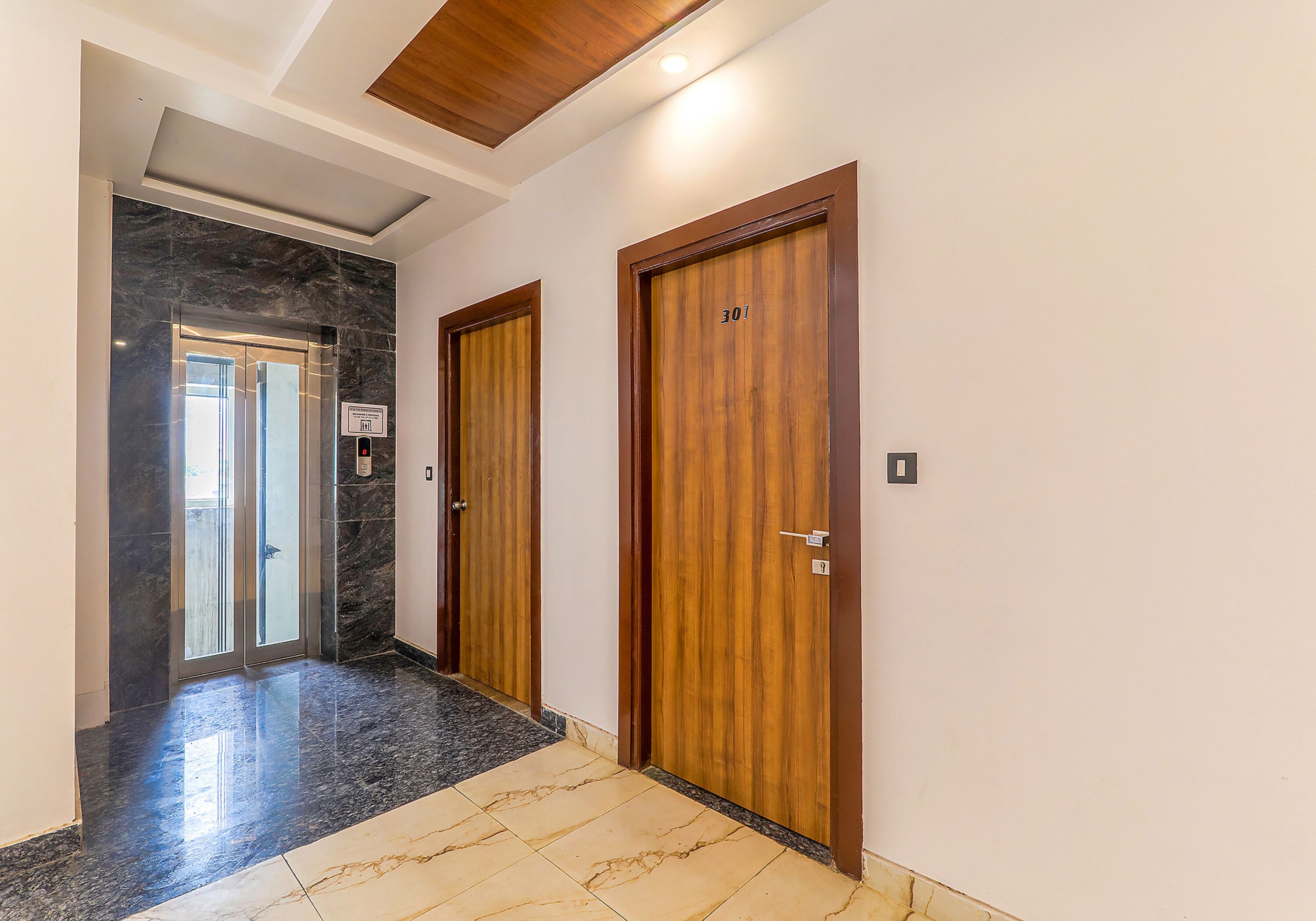 A hallway with two wooden doors, a glass elevator, and a marble floor.