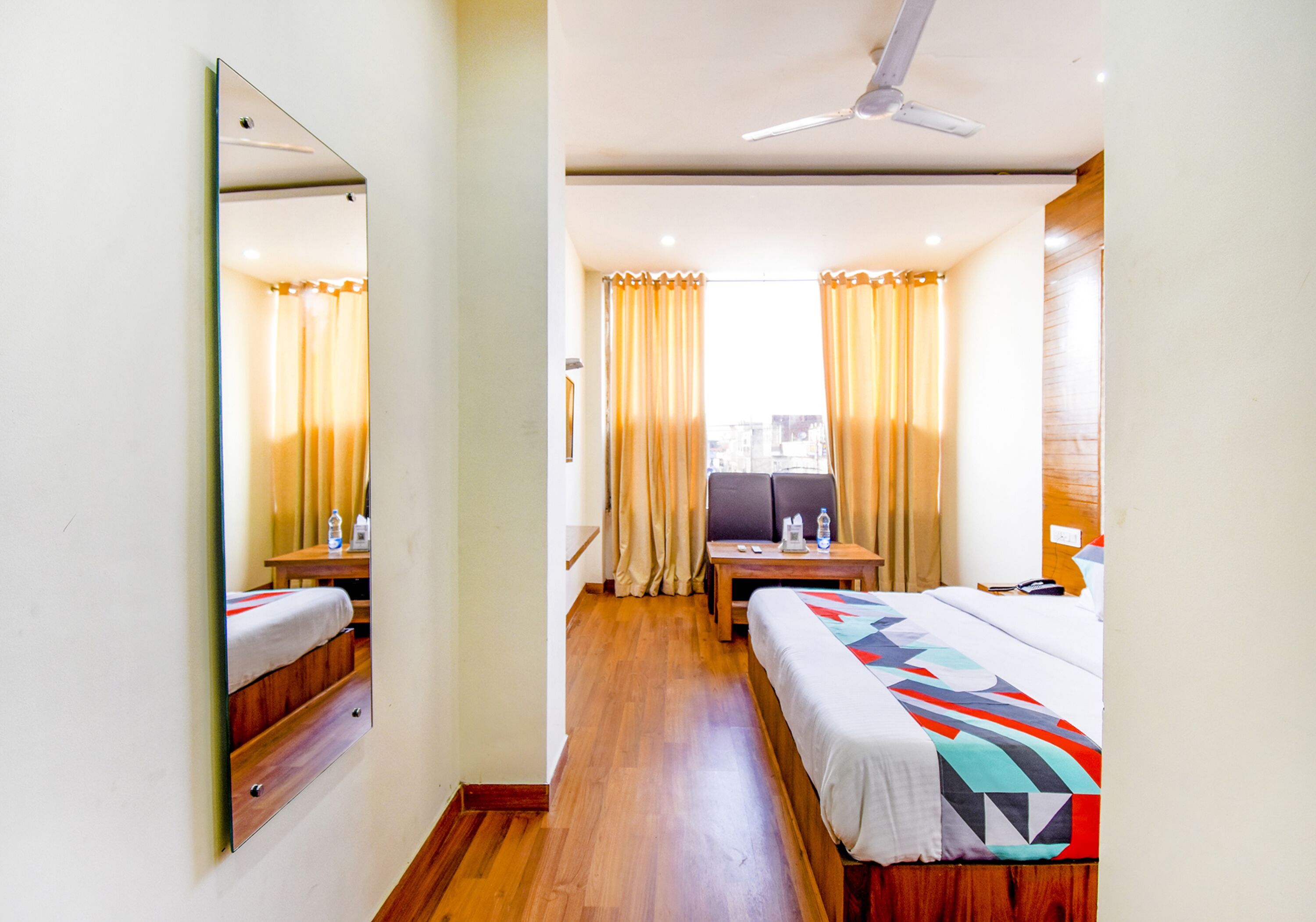 A hotel room with a bed, a desk, a chair, a mirror, and a ceiling fan.