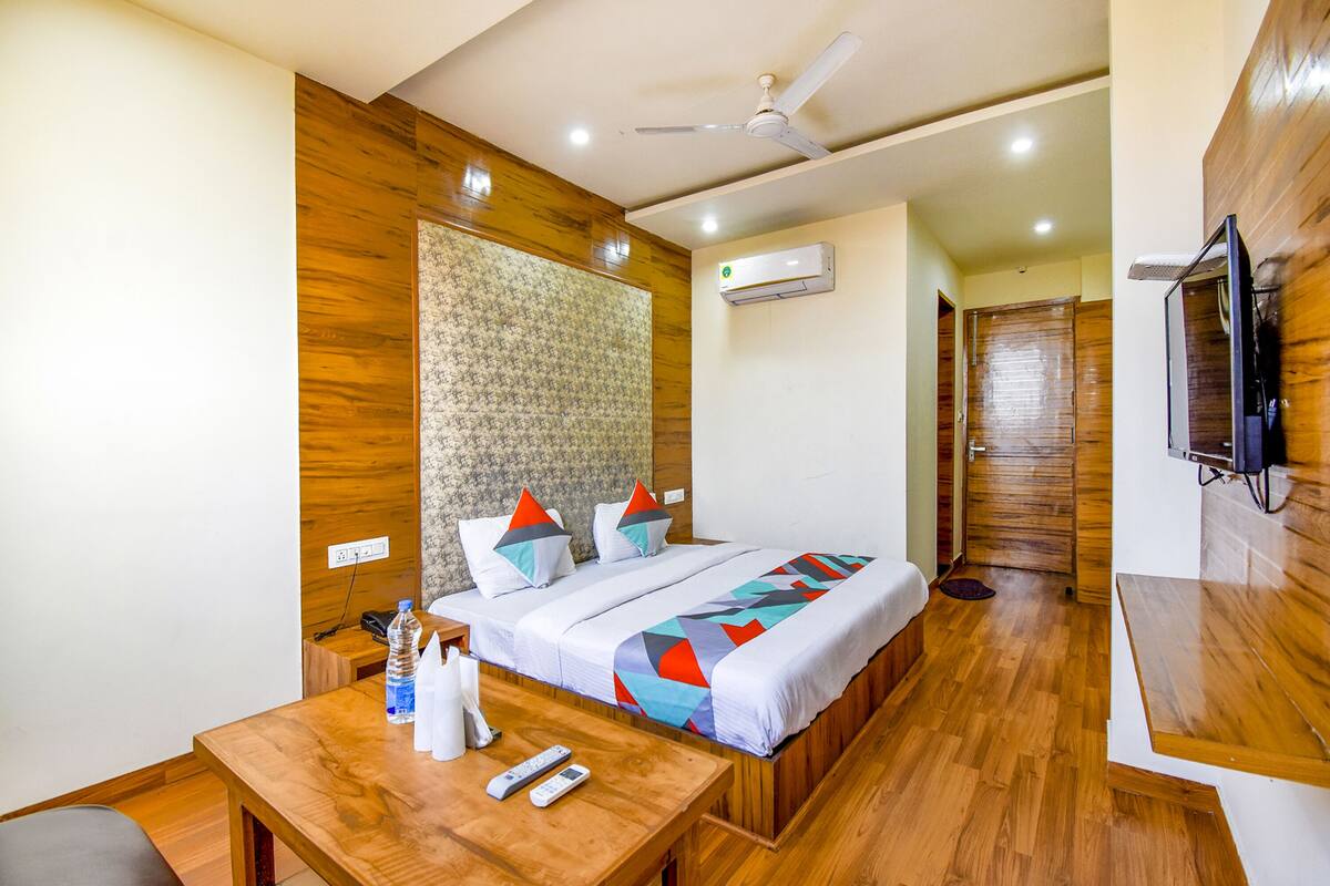 Habitación de hotel con piso de madera, una cama con almohadas de colores, un escritorio de madera con una botella de agua, un control remoto y un televisor montado en la pared.
