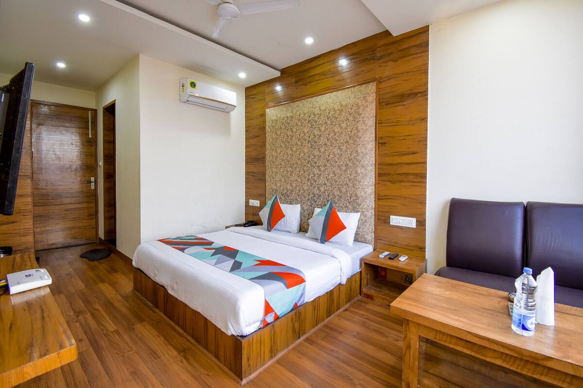 Habitación de hotel con piso de madera, una cama con ropa de cama colorida, una mesita de noche, una mesa con una botella de agua, un sofá y un ventilador de techo.