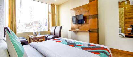 A hotel room with a bed, a desk, a chair, a TV mounted on the wall, and a window with curtains.