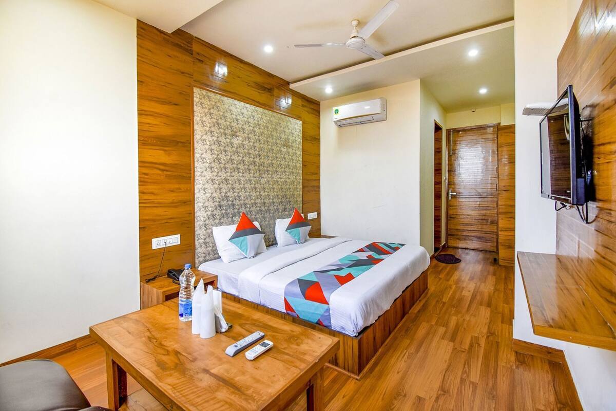 Una habitación de hotel con piso de madera, una cama con colcha de diseño geométrico y colores vivos, cabecera de madera, ventilador de techo, unidad de aire acondicionado, televisor montado en la pared y una pequeña mesa de madera con un control remoto y una botella de agua.