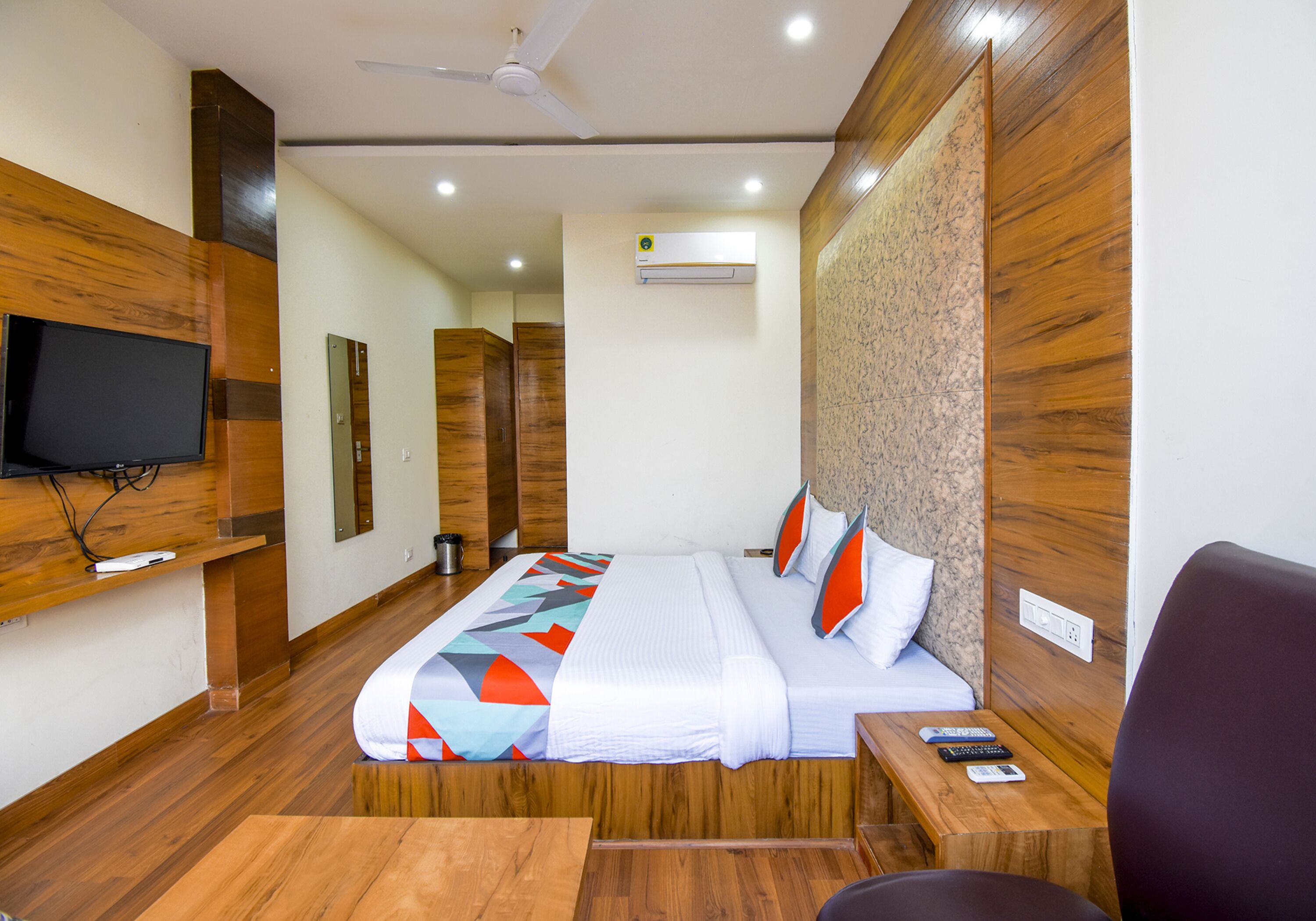 A hotel room with a wooden floor, a bed with a colorful bedspread, a TV mounted on the wall, and a ceiling fan.