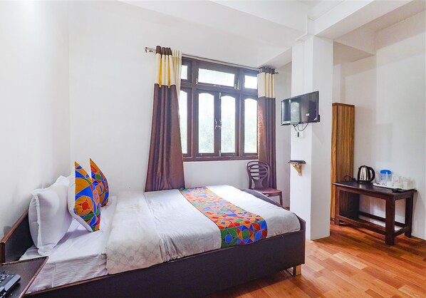 A hotel room with a bed, a TV mounted on the wall, a wooden table with a kettle and cups, and a window with curtains. - Fabexpress Laxmi Niwas (Gangtok)