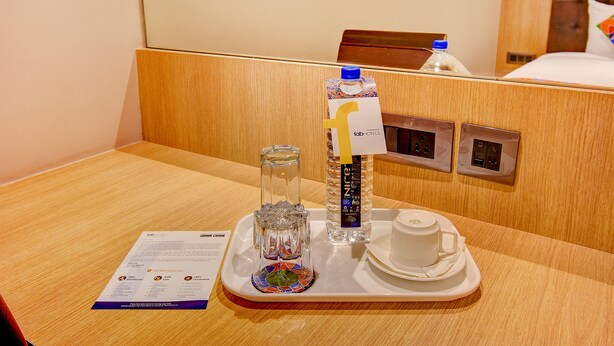 A hotel room with a wooden headboard, a bedside table with a tray containing a glass, a bottle of water, and a cup.