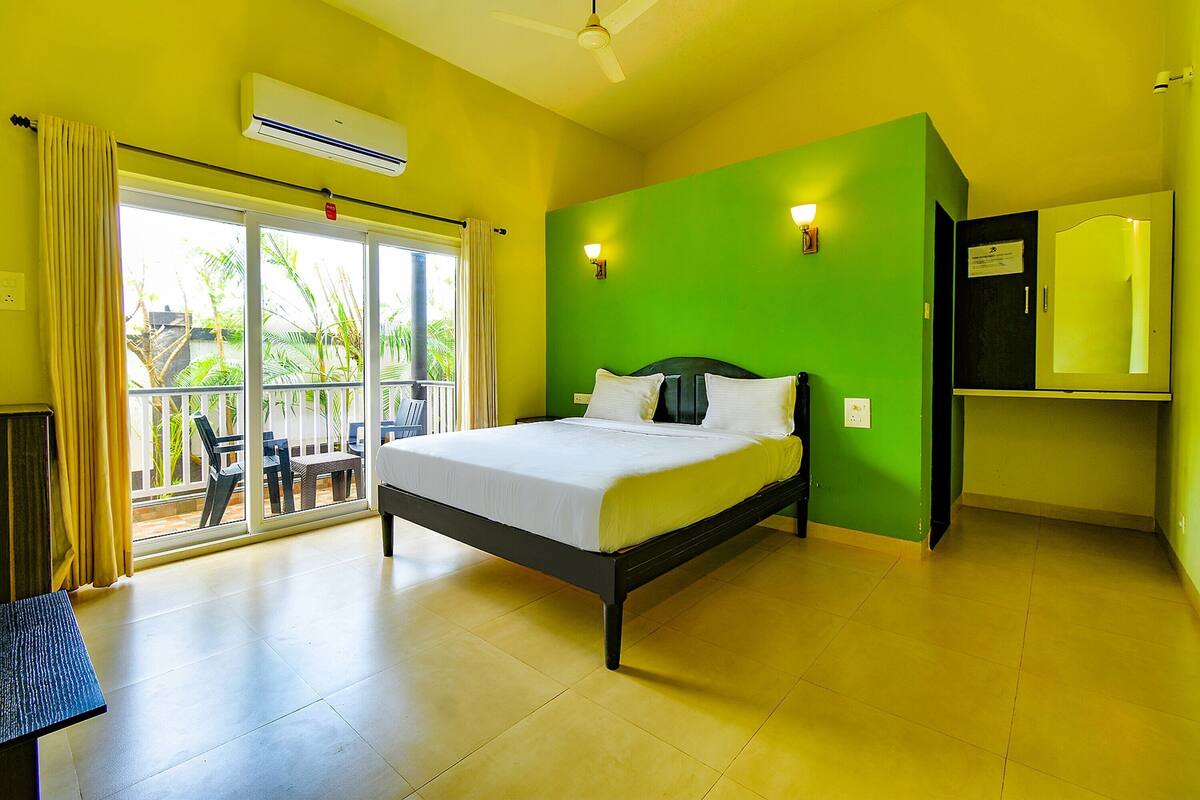 A room with a bed, a ceiling fan, an air conditioning unit, a sliding glass door leading to an outdoor area with chairs and a table, and a small shelf with a mirror.