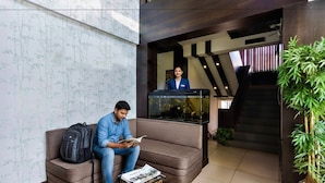 Un hall d’entrée moderne avec un comptoir d’accueil, un escalier et un espace détente où une personne est assise, en train de lire un magazine.